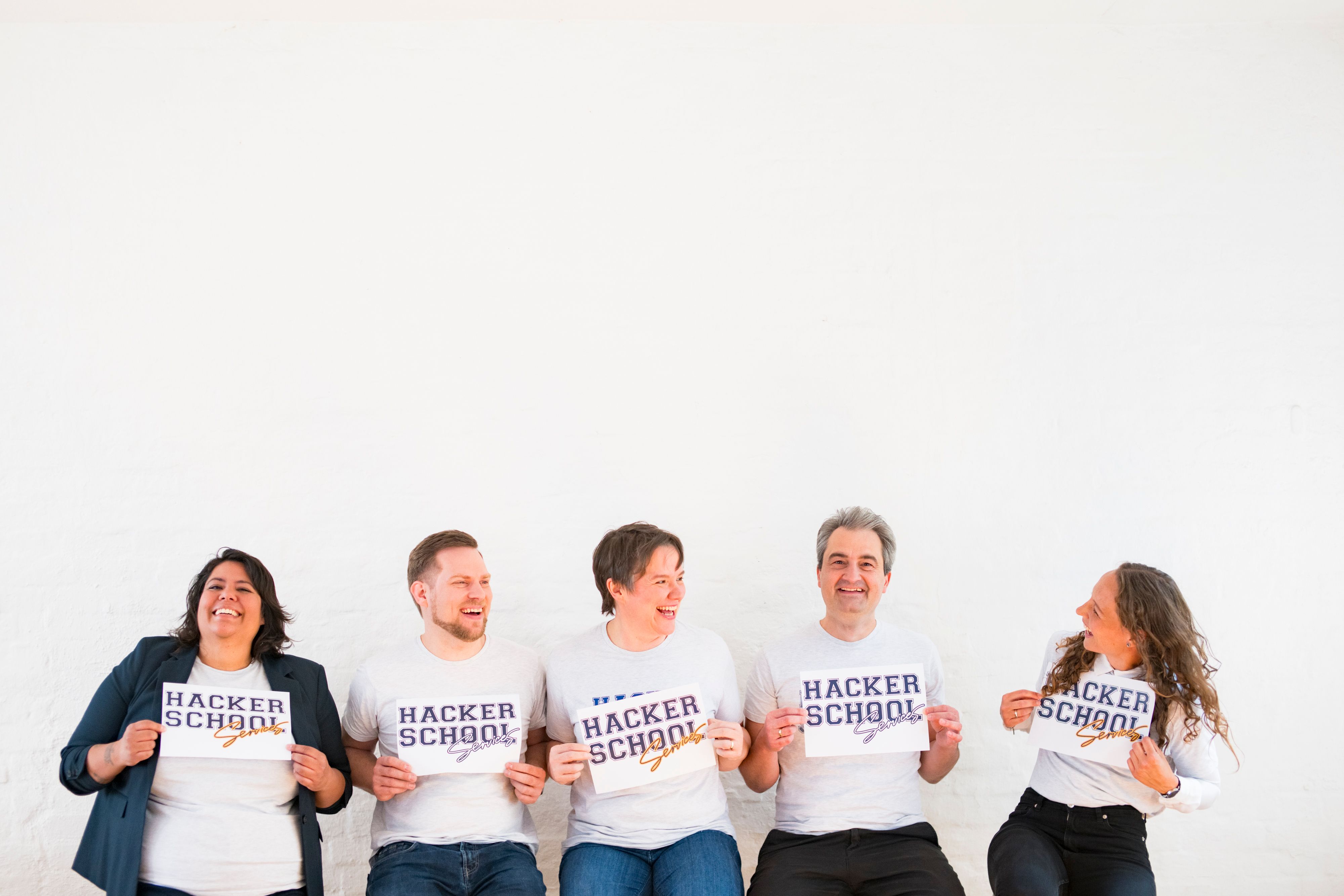 Die fünf Gründer:innen der Hacker School Services GmbH nebeneinander vor einer weißen Wand, lachend, mit Schildern in der Hand
